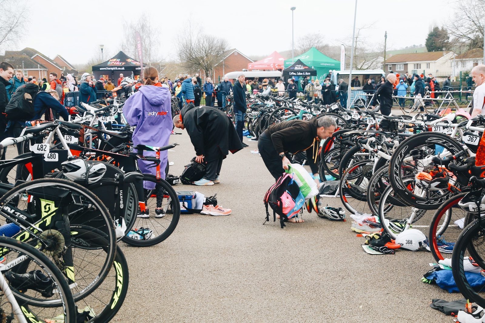 Athletes prepare their gear in a crowded bicycle transition area during a triathlon event. They organize equipment and change clothes among numerous parked bikes, with spectators and other participants in the background on a slightly overcast day.