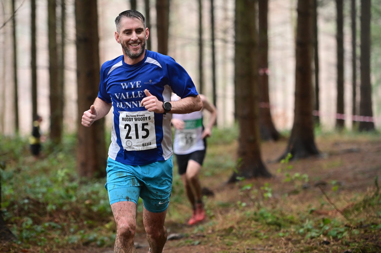 A man with a numbered bib 215 on his shirt, participating in a forest trail run, smiles and gives two thumbs up. He wears a blue "Wye Valley Runners" shirt and blue shorts. Another runner is in the background, partially obscured by a tree.