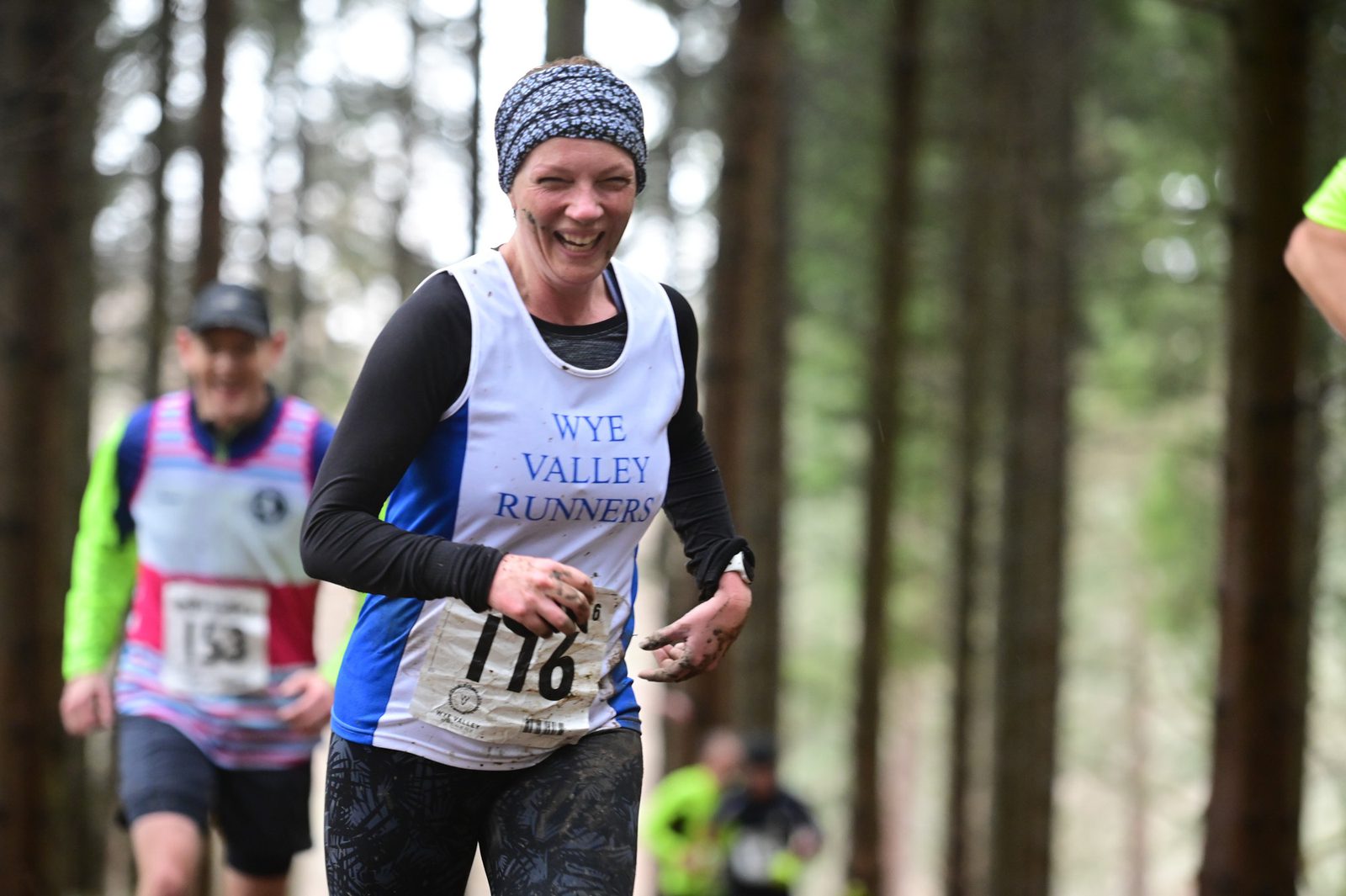A woman, wearing a Wye Valley Runners jersey and a knit cap, smiles as she runs through a forested trail. Her race bib number is 116. Other runners in brightly colored jerseys are visible in the background, and the path is surrounded by tall trees.