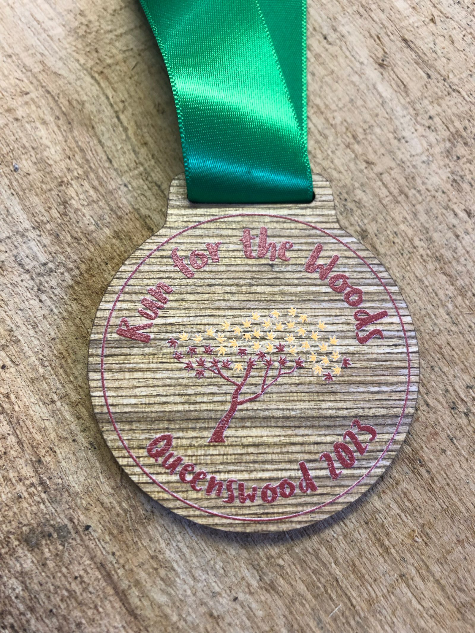 A wooden medal with a green ribbon is engraved with "Run for the Woods" at the top and "Queenswood 2023" at the bottom. In the center, there is a tree design surrounded by small yellow stars. The medal lies on a wooden surface.