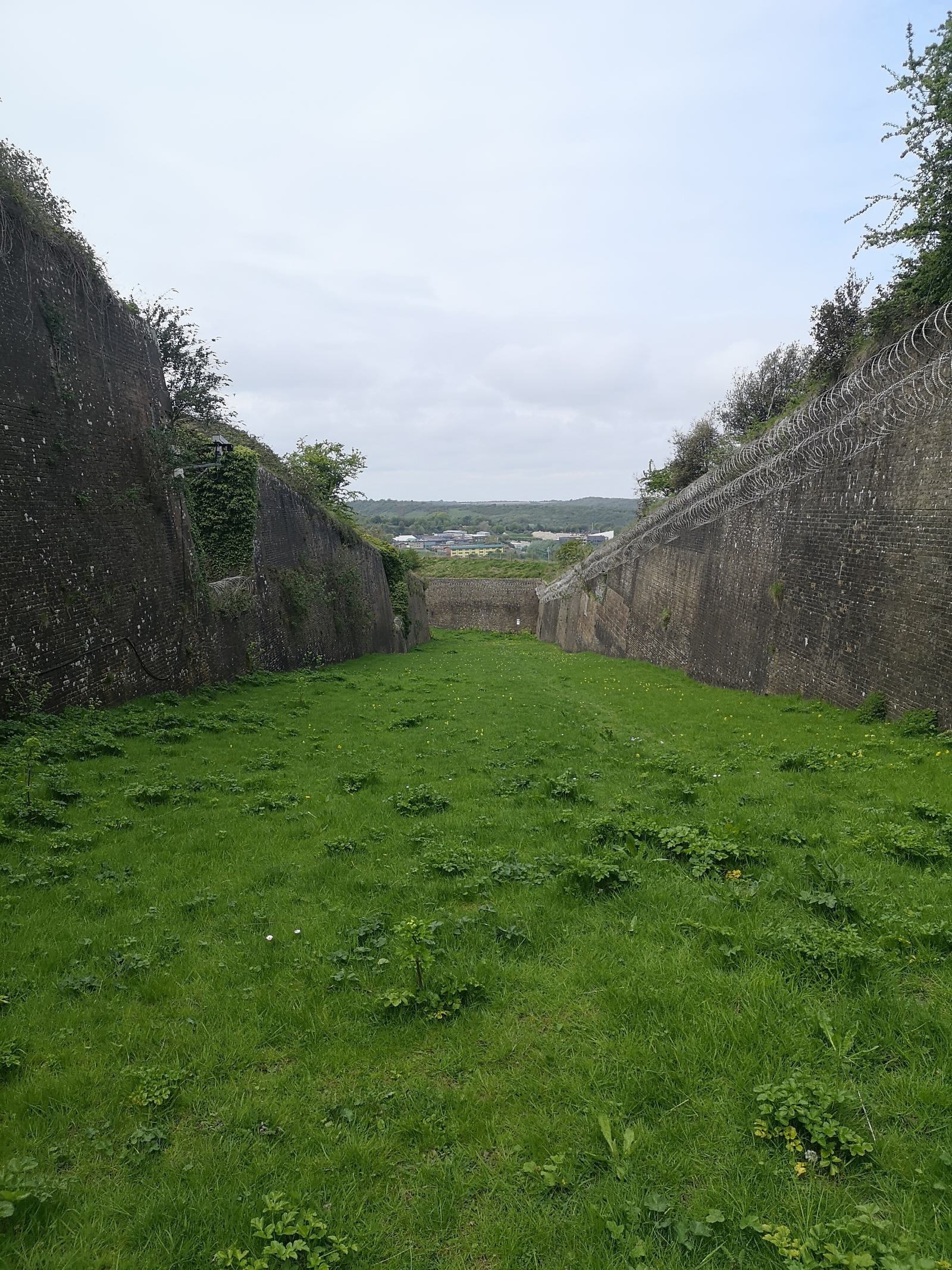 A grassy pathway between two tall, weathered stone walls lined with barbed wire, leading down to a distant view of a town and hills under a cloudy sky. The pathway is slightly overgrown with patches of grass and small plants.