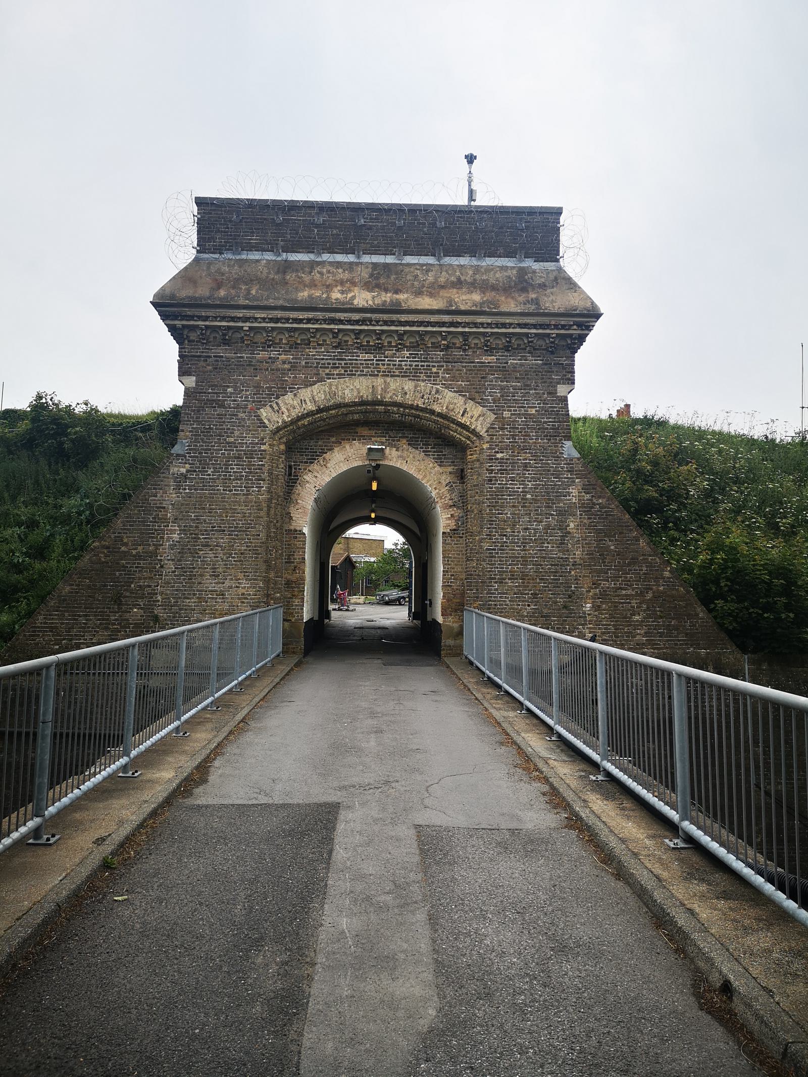 A historical brick arch bridge over a road with a metal fence on both sides. The bridge has an arched opening through which a road can be seen, and it is topped with a layer of barbed wire. In the background, green foliage and a cloudy sky are visible.