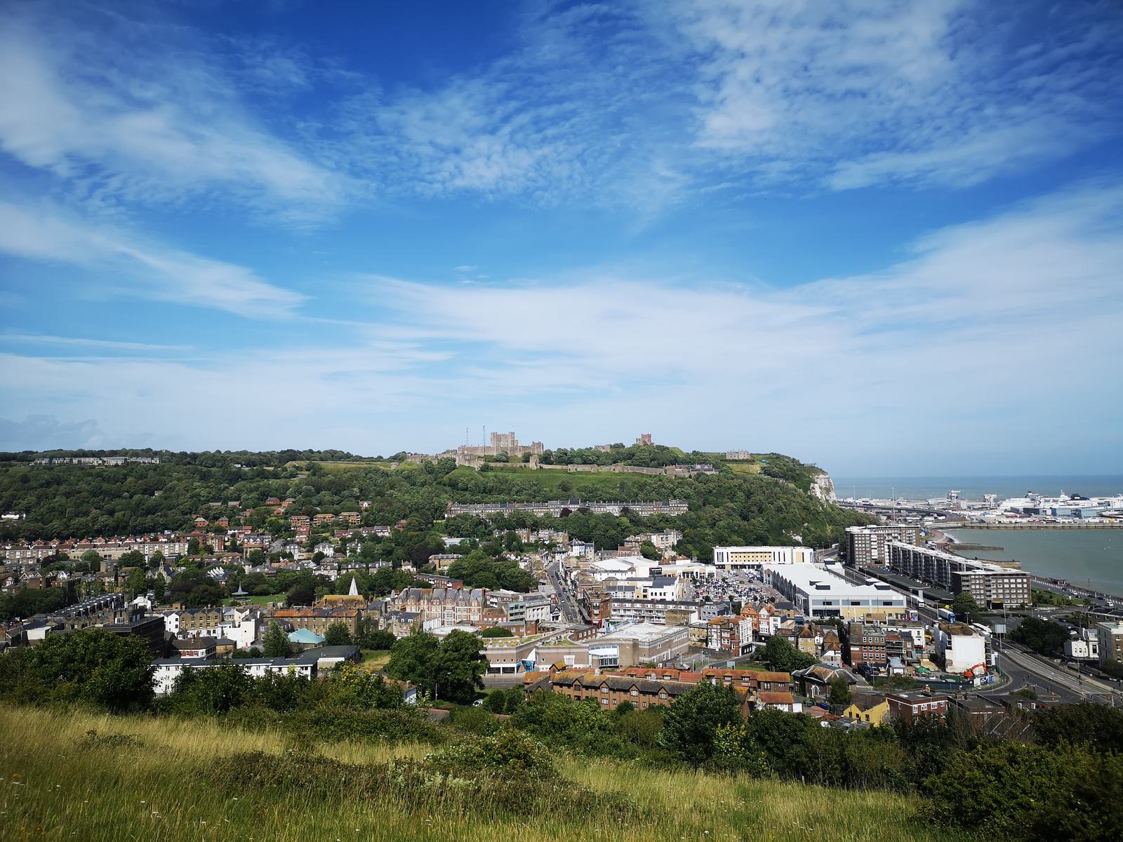 A scenic view of a town by the coast under a blue sky with scattered clouds. The town's buildings spread out along the shoreline and up a green hill, where a large castle is prominently situated at the top. In the background, the sea extends to the horizon.