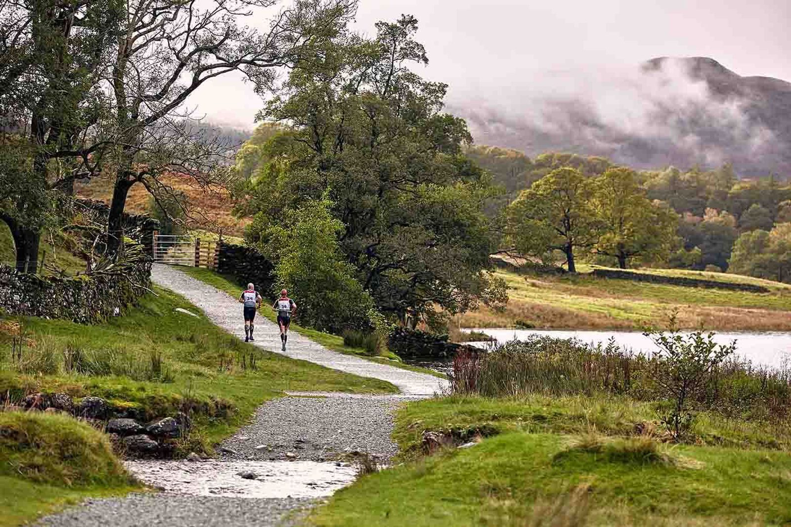 Two cyclists ride on a winding gravel path through a lush, green landscape by a calm lake. Misty mountains and trees surround them, and a stone wall runs parallel to part of the path. The scene is serene and overcast.