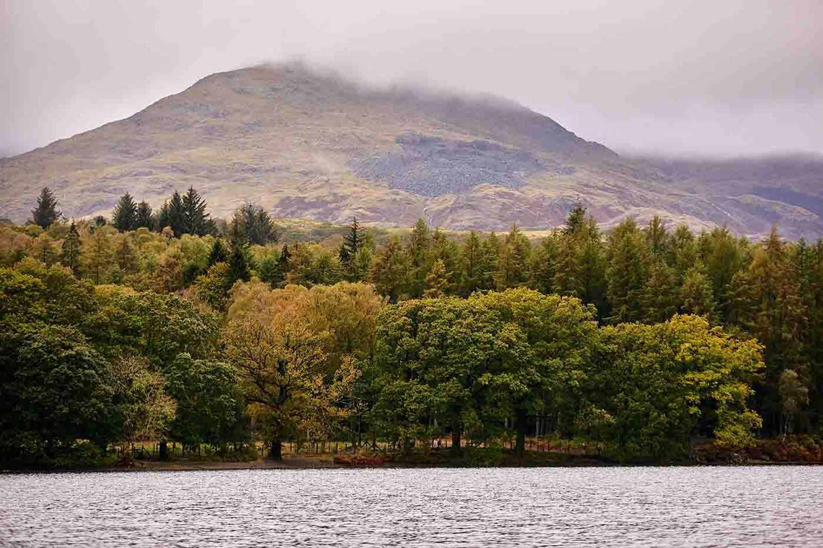 A mist-shrouded hill rises above a dense forest of autumn trees with changing leaves, ranging from green to golden yellow. The calm waters of a lake in the foreground reflect parts of the scenery under a cloudy sky.