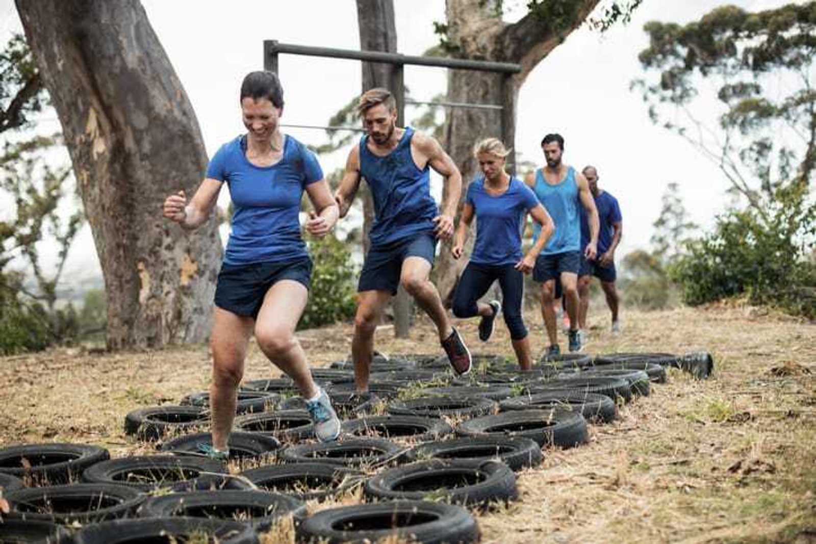A group of five people, dressed in blue shirts and black shorts, are outdoors engaged in a tire workout exercise. They are running and stepping through a series of tires placed on the ground. Trees and greenery are visible in the background.
