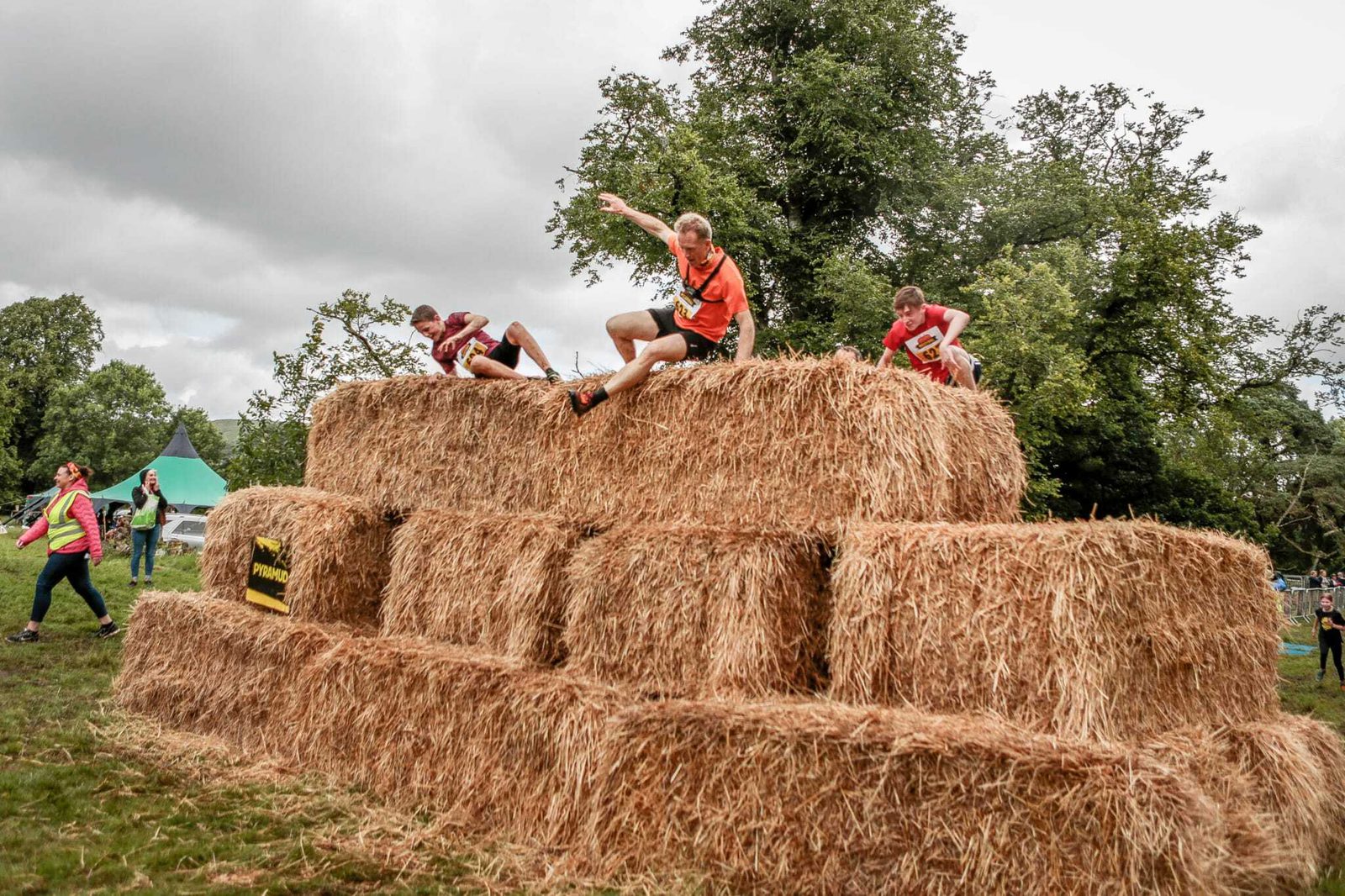 Participants climb and jump over a large stack of hay bales in an outdoor obstacle course race. Trees and a tent can be seen in the background, while spectators and event staff watch. The scene is cloudy, giving the event an adventurous and challenging atmosphere.