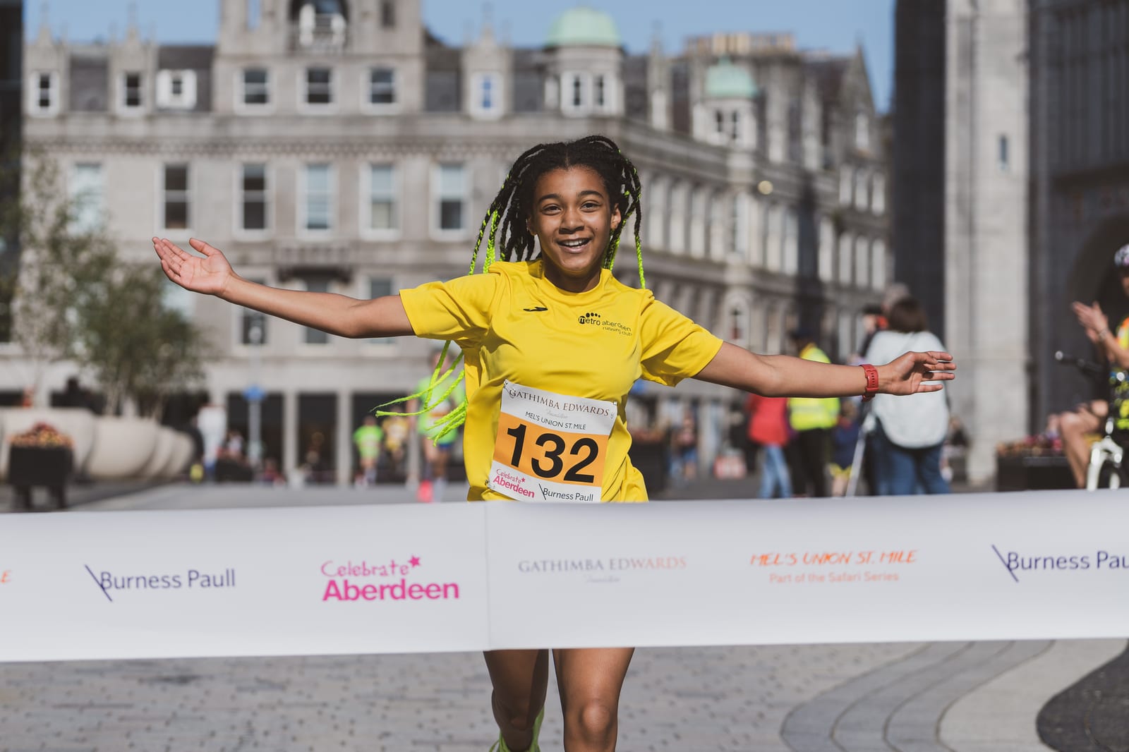 A young girl wearing a yellow shirt and race number 132 smiles joyfully with arms outstretched as she crosses the finish line of a city marathon event. City buildings and other participants are seen in the background.