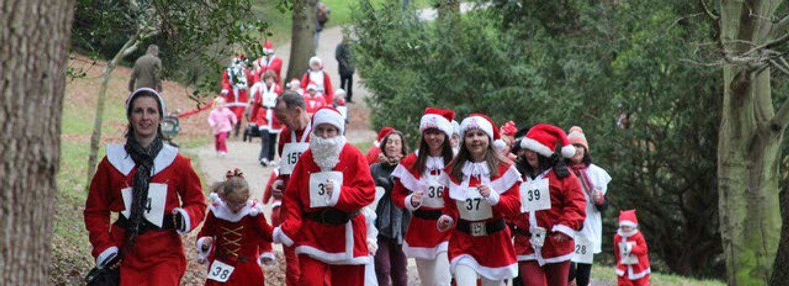 A group of people dressed in Santa Claus costumes are jogging on a tree-lined path, participating in a festive fun run. Some are wearing numbered bibs, and there are both adults and children involved, all enjoying the outdoor event.