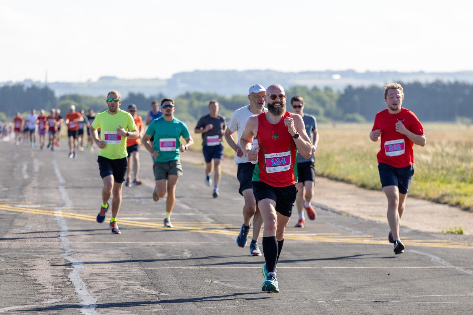 A group of runners participating in a race on a paved road, with numbers pinned to their shirts. The scene is set outdoors on a clear day, with a backdrop of fields and distant trees under a bright sky.