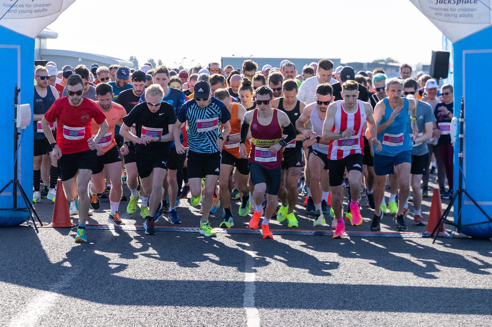 A large group of runners in athletic gear start a race on a sunny day. They are all gathered closely together as they cross the starting line, with several wearing brightly colored shoes.