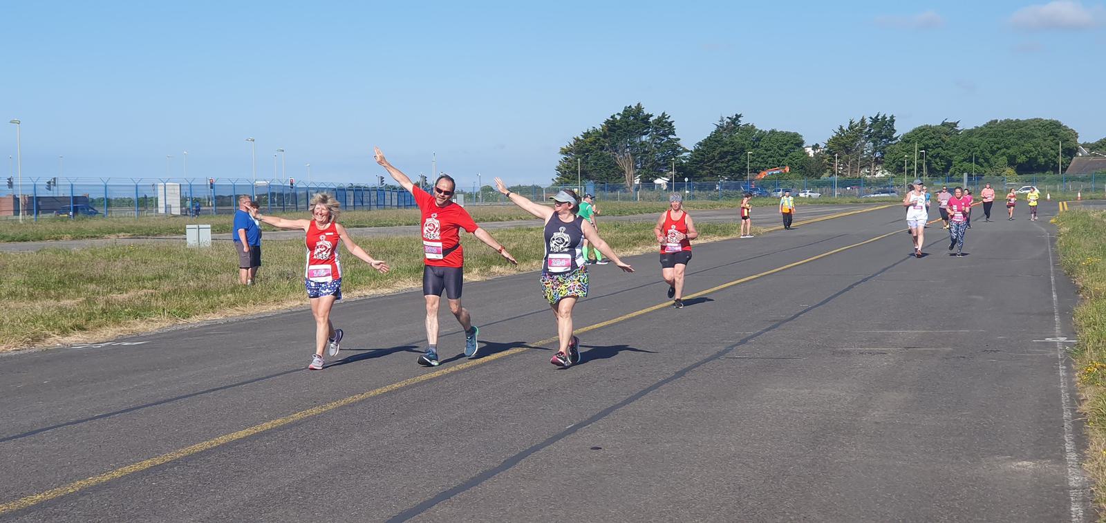 A group of runners in colorful attire are happily waving as they jog down a sunny, open road. The scene is set in a rural area, with clear skies and distant trees in the background. Other runners are visible along the road ahead and behind.