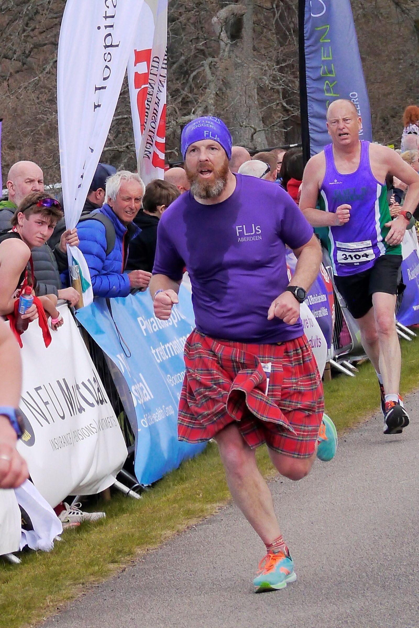 A man with a beard, wearing a purple shirt, a flannel kilt, and a purple headband, is running in a race. Behind him, another male runner in purple and green athletic gear is also participating. Spectators in jackets are watching and cheering from behind a barrier.