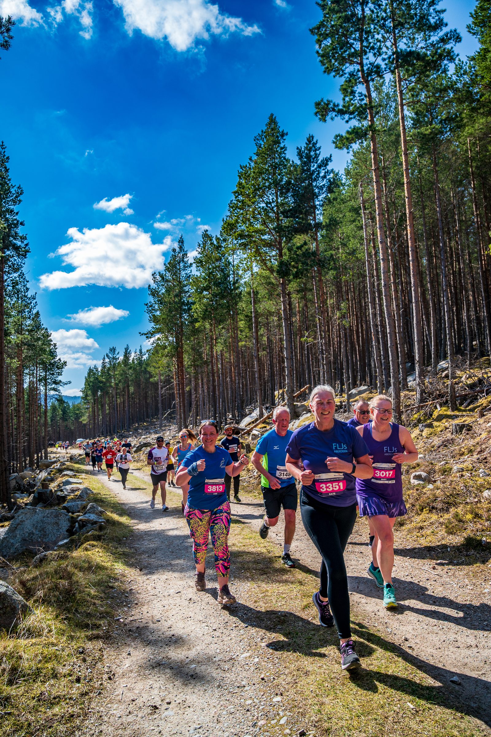 A group of runners participate in a trail race through a forest under a bright blue sky with scattered clouds. They are on a dirt path lined with trees and rocks. Some runners are smiling as they move forward, all wearing colorful race bibs and athletic attire.