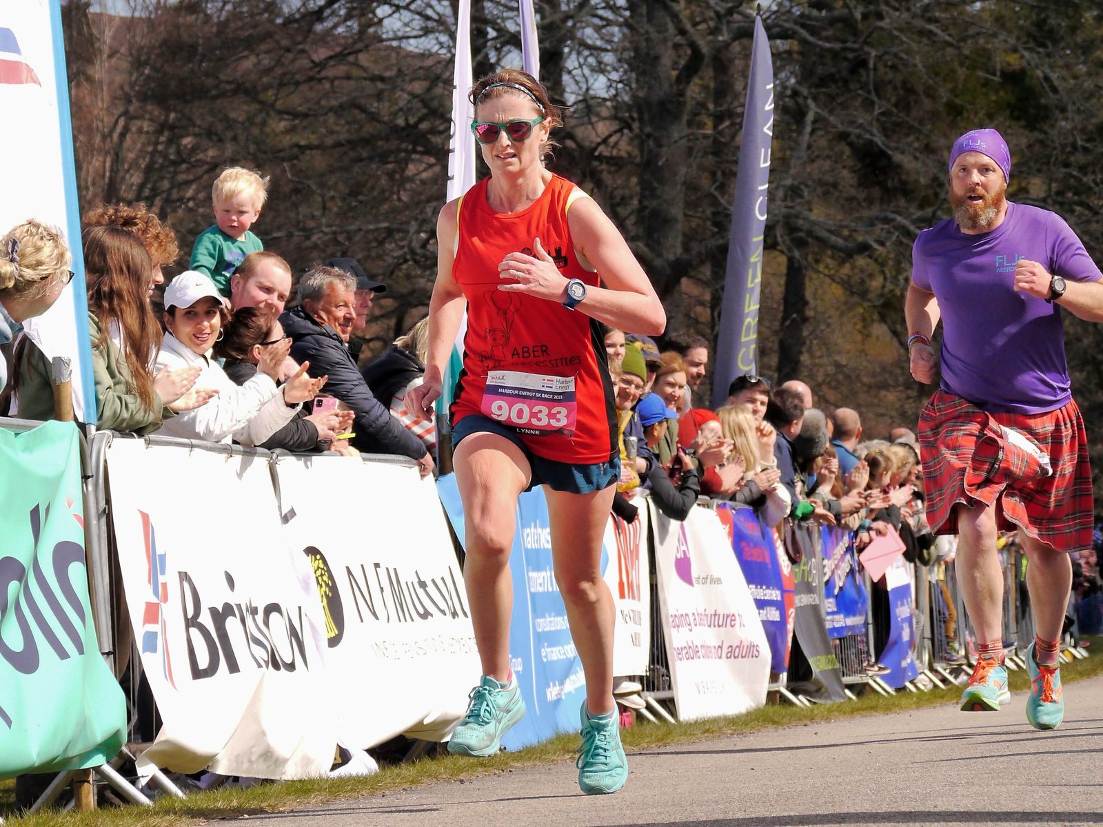 A woman in a red tank top and blue shorts is running in a race, wearing bib number 9033. Crowds of spectators line the race route behind barriers, cheering the runners. A man in a purple shirt and plaid kilt is running behind her. Trees and banners are visible in the background.