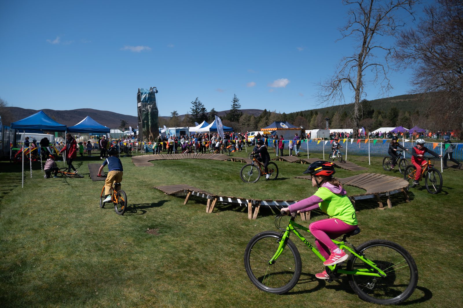 Children and adults ride bicycles on a wooden track in a grassy area during an outdoor event. Tents and colorful flags are set up in the background, with a crowd of spectators gathering. A large statue and trees are visible under a clear blue sky.