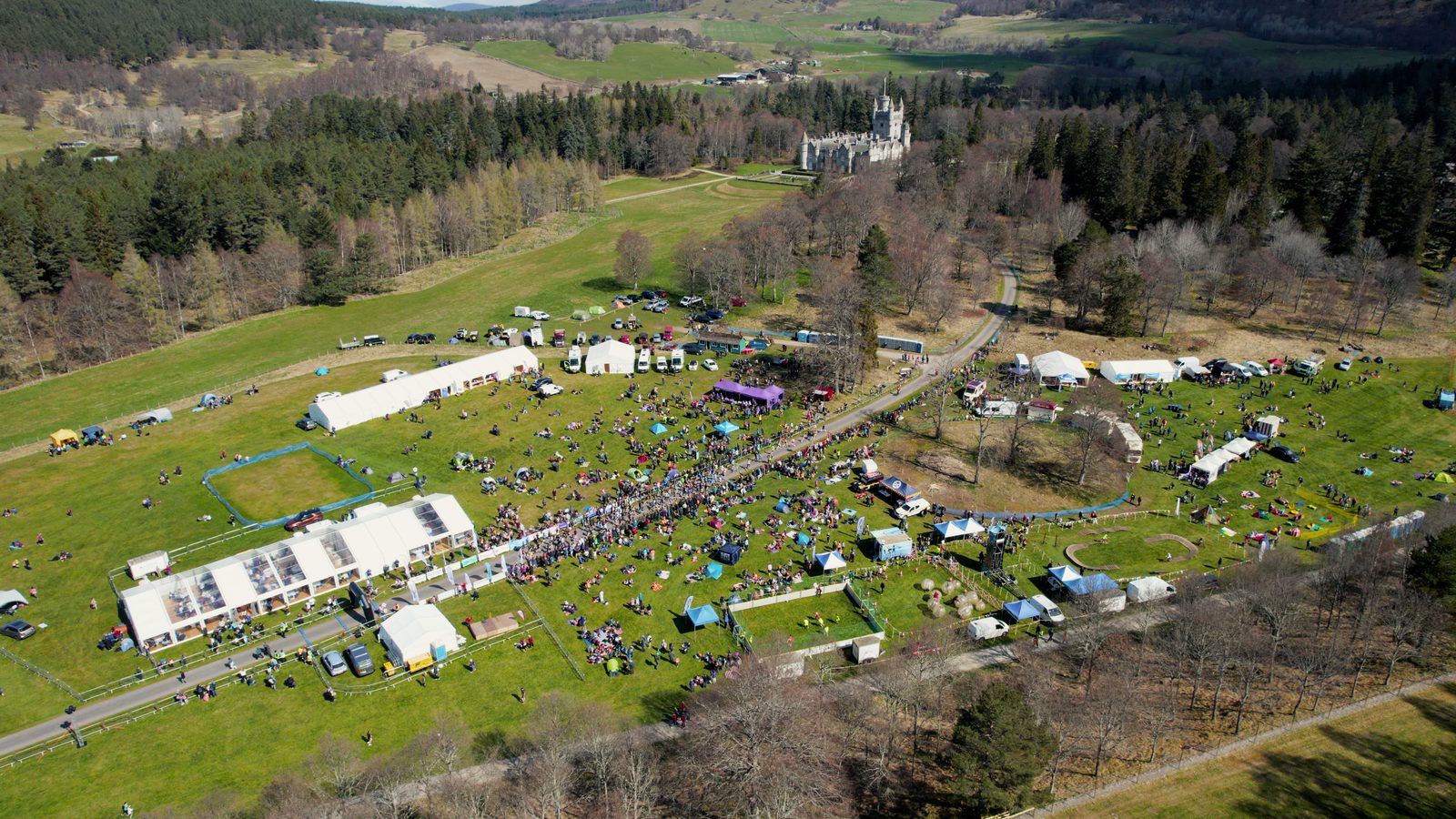 Aerial view of an outdoor festival on a sunny day, with numerous white tents and crowds of people gathering on a grassy field. Surrounding the field are trees and a lightly forested area, with a grand building visible in the background.