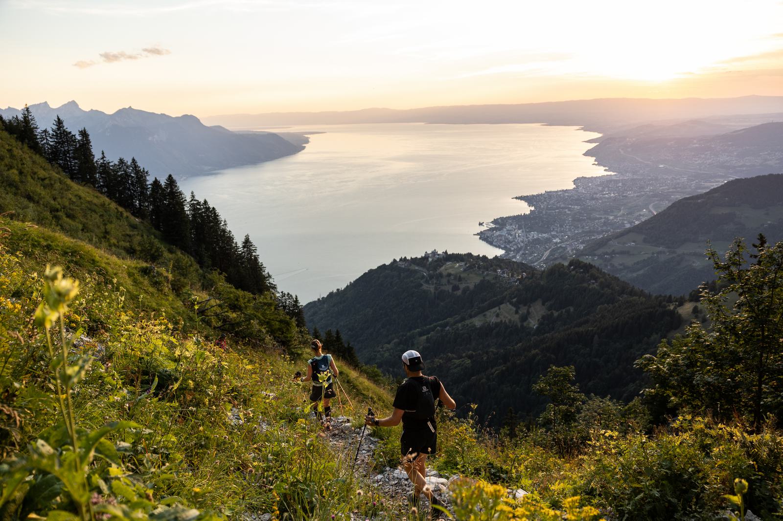 Two people are trail running on a lush, green hillside path, overlooking a breathtaking landscape featuring a large body of water, mountains, and a setting sun casting a golden glow over the scene. The path is surrounded by wildflowers and greenery.