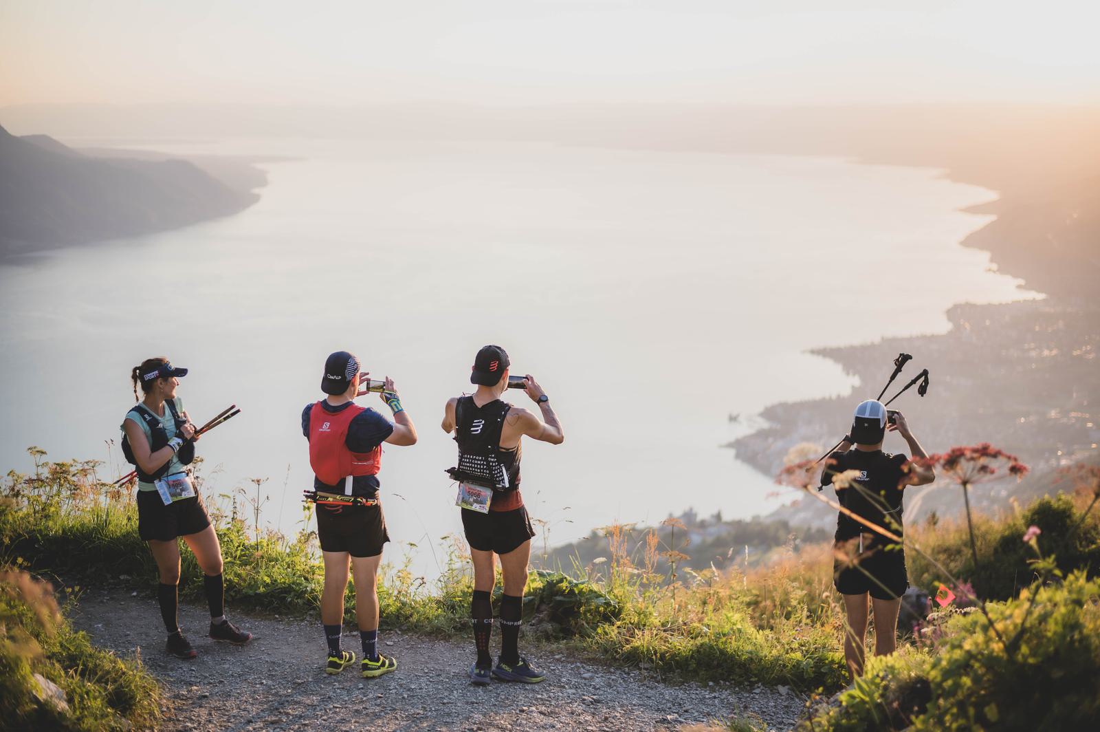 Four people are standing on a scenic mountaintop trail overlooking a large body of water at sunset. They are dressed in running gear and are taking photos of the view, with hiking poles in hand. The sky is hazy and the overall atmosphere is serene and peaceful.