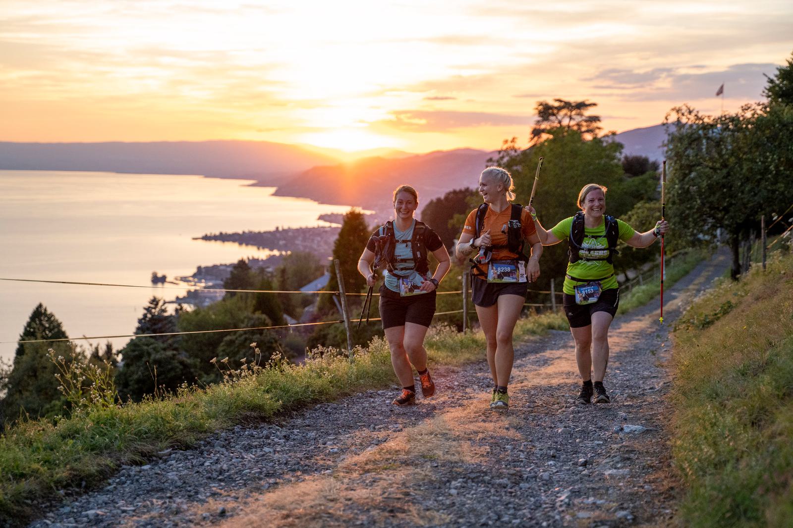 Three runners are trekking on a gravel path overlooking a lake at sunset. They smile and converse as they move, with numbers pinned to their attire indicating participation in an organized event. The sun sets on the horizon, casting a warm glow over the landscape.