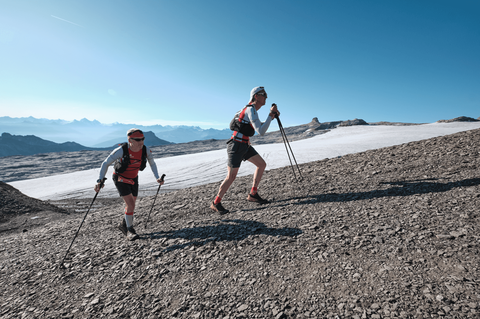 Two people with trekking poles hike up a rocky mountain slope under a clear blue sky, with snow and distant mountain peaks visible in the background.