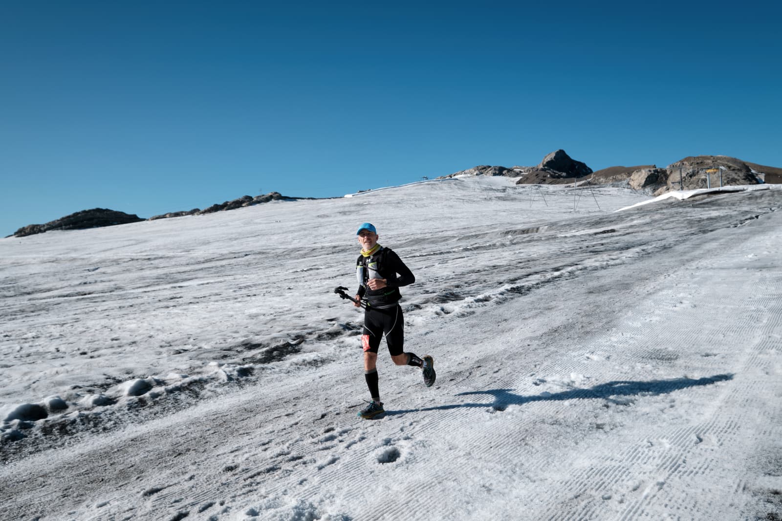 A person runs on a snowy, sloped landscape under a clear blue sky, wearing athletic gear and holding trekking poles. Rocky peaks rise in the background.