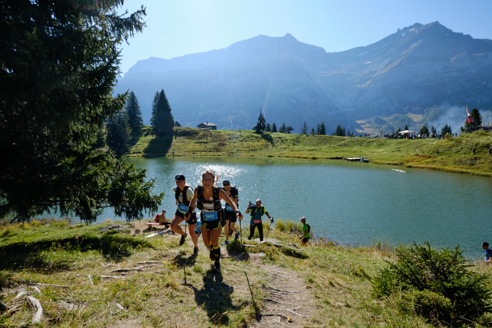 Runners in athletic gear ascend a grassy trail beside a mountain lake, surrounded by pine trees and mountains under a clear blue sky, during a trail race or marathon event.