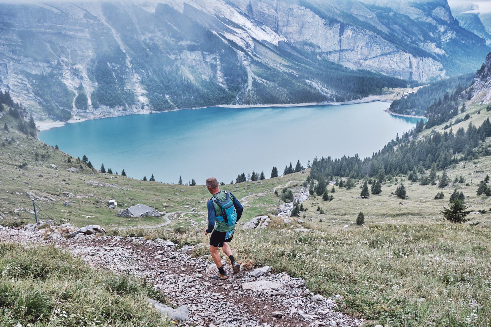 A person with a backpack hikes down a rocky trail towards a scenic, turquoise lake surrounded by mountains. The landscape is dotted with trees and patches of grass, and the sky is overcast, casting a soft light on the natural scenery.