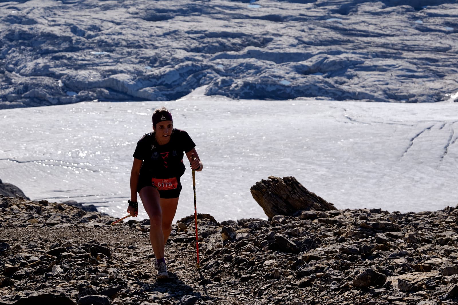 A person with trekking poles hikes uphill on a rocky trail, with a large glacier or snowy landscape in the background under bright daylight.