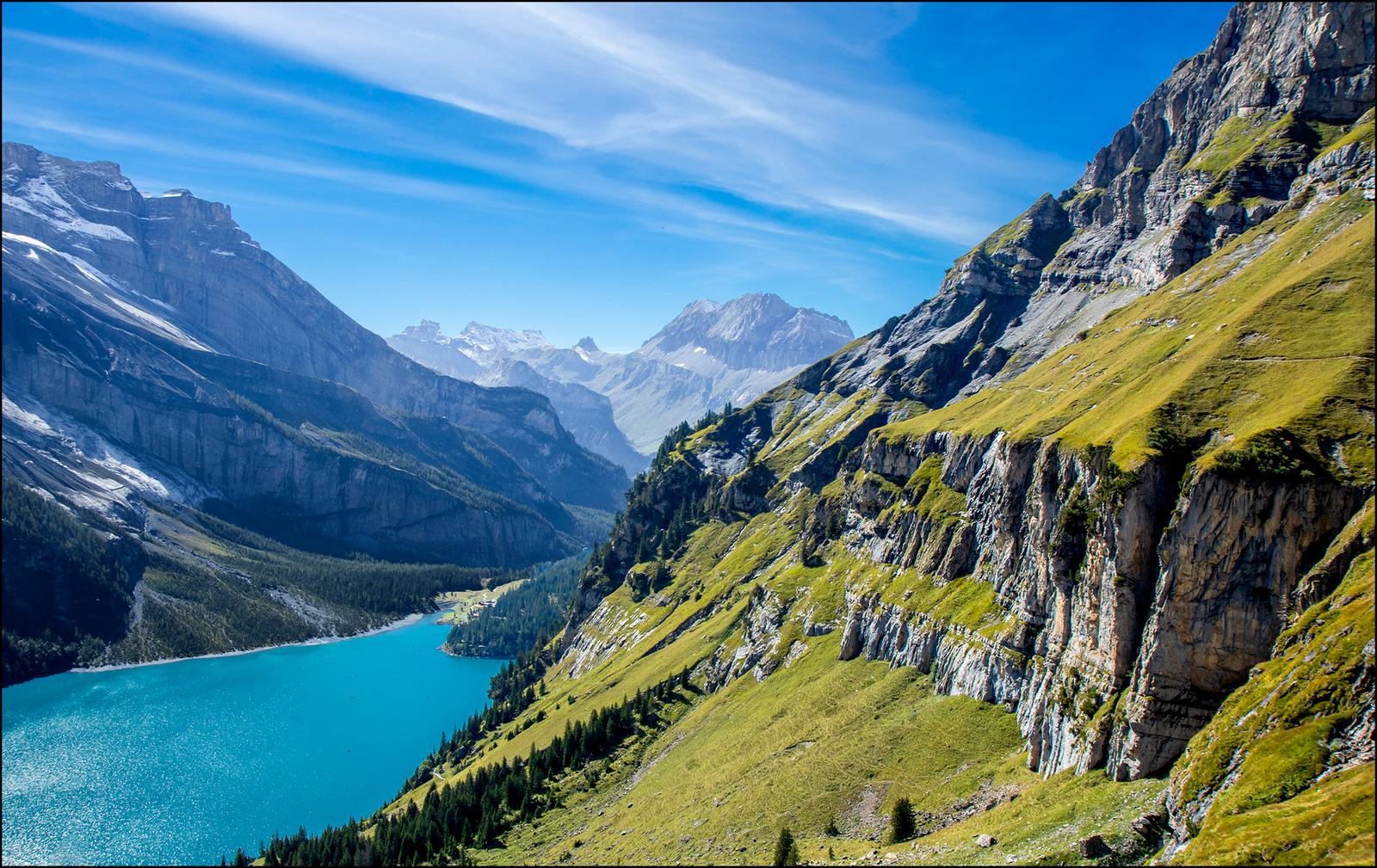 A pristine blue lake is nestled amongst mountainous terrain with green slopes and rocky cliffs. The clear sky is a vibrant blue, casting light across the landscape. Snow-capped peaks are visible in the distance, contributing to the panoramic alpine scenery.