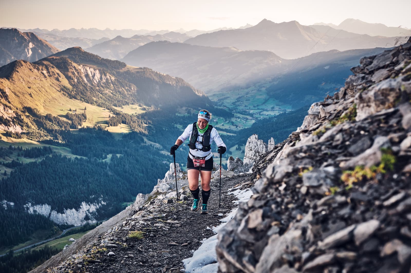 A runner with trekking poles ascends a rocky mountain trail, surrounded by dramatic peaks and a scenic valley below, during daylight in an alpine landscape.