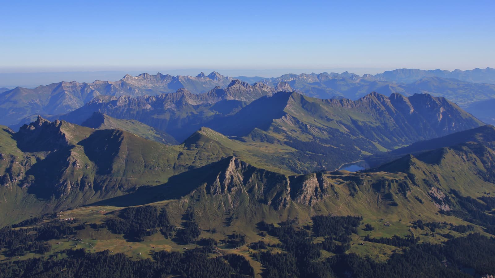 A panoramic view of green, rugged mountain peaks under a clear blue sky, with distant ridges fading into the horizon. Sunlight highlights the contours and valleys of the landscape.