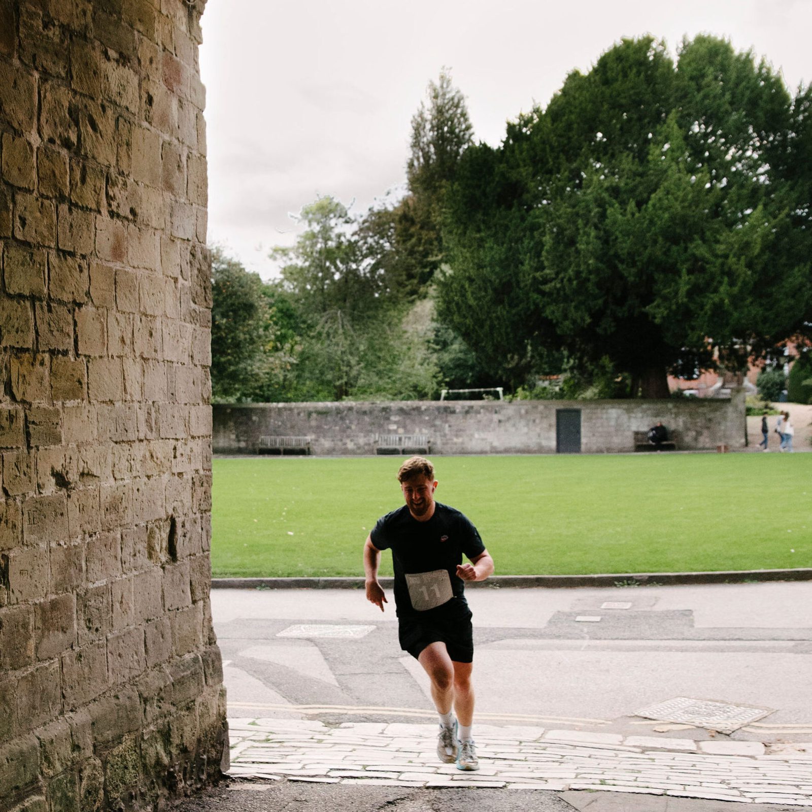A runner in a black shirt and shorts jogs through an old stone archway on a paved path, with green grass and trees visible in the background. A hanging lantern is attached to the top of the arch. The scene is overcast.