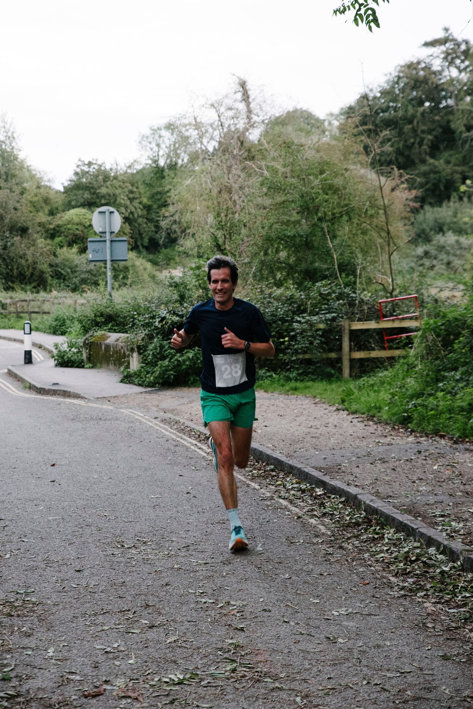 A man is running along a paved path in a park. He is wearing a black t-shirt, green shorts, and running shoes. He holds his two thumbs up while smiling. The background has greenery, including trees and bushes, and a road sign is seen in the distance.