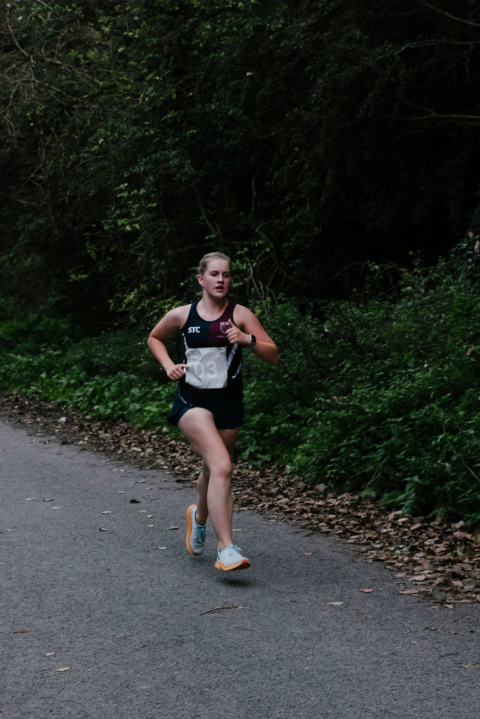 A person running on a paved path surrounded by dense greenery. They are wearing a black athletic outfit with a visible race number pinned to their chest. Leaves are scattered on the ground, suggesting an autumn setting.
