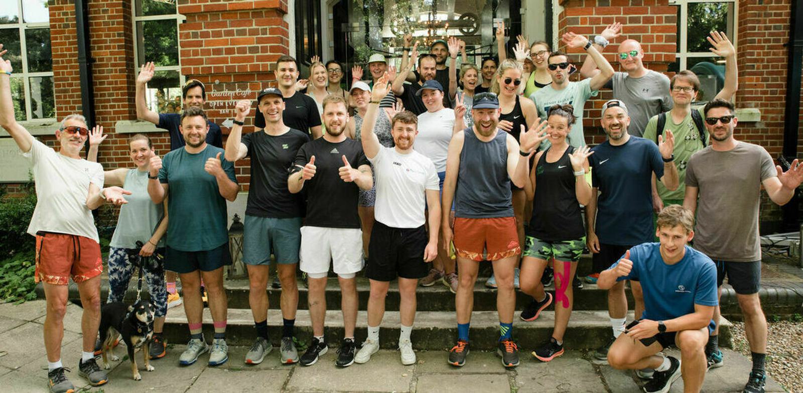 A large group of people in athletic wear stand in front of a brick building, smiling and raising their hands in celebration. Some are giving thumbs up. The setting is outdoors, and there's greenery visible around them.