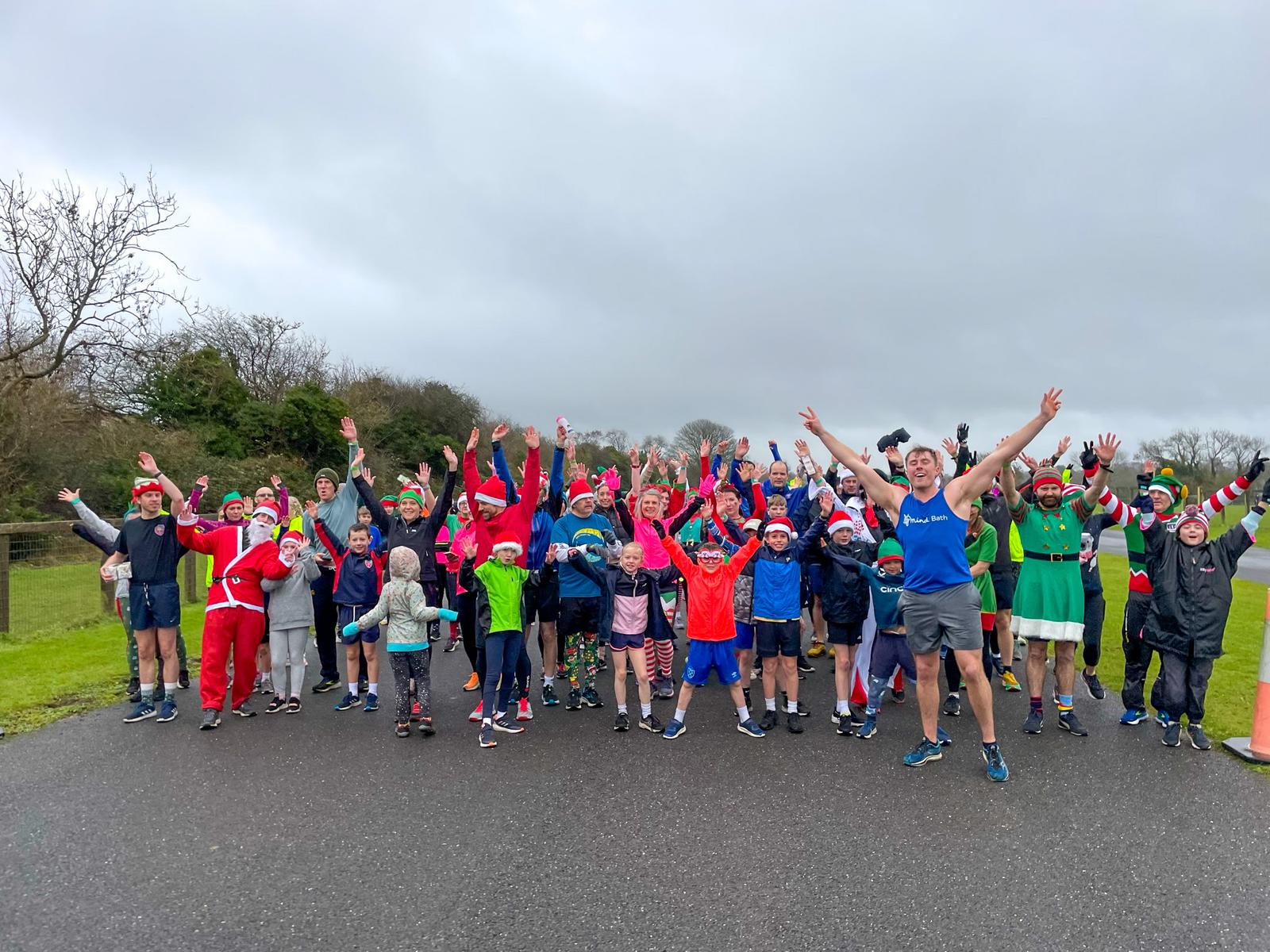 A group of people dressed in festive and colorful holiday attire, including Santa hats and elf costumes, pose with their arms raised in excitement during a fun run event held on a cloudy day. The participants are gathered on a paved path with trees in the background.