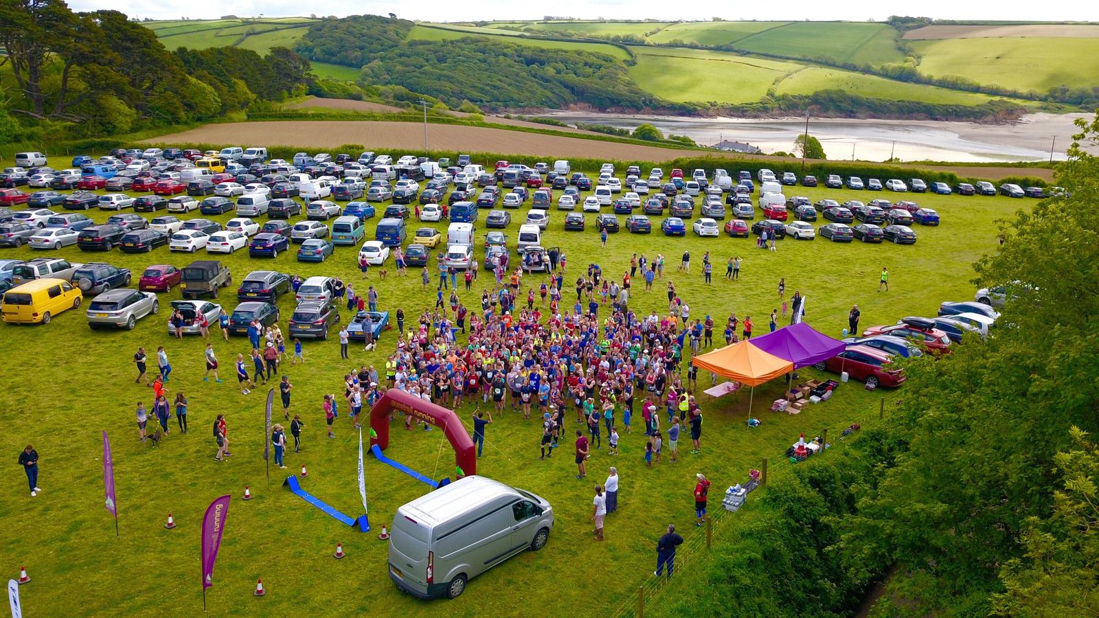 Aerial view of a large group of people gathered on a grassy field for an outdoor event. Numerous parked cars fill the background. Two tents and a van are visible near the crowd. The landscape features lush greenery and distant rolling hills.