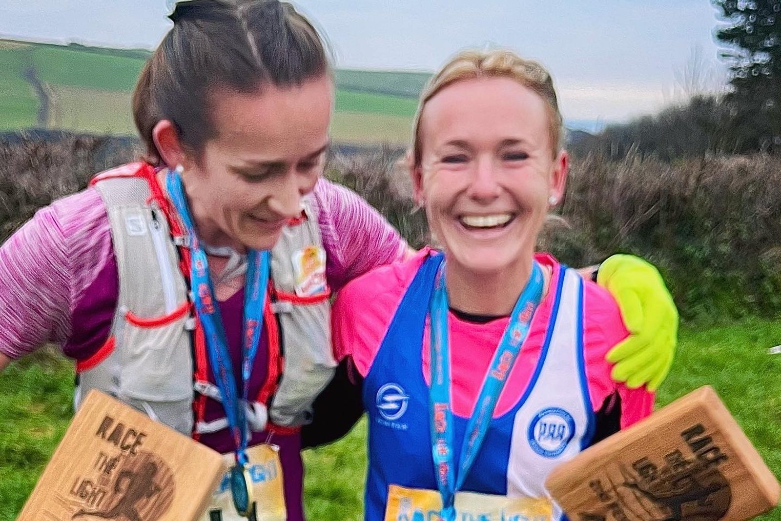 Two runners are smiling and holding wooden plaques with "RACE THE LIGHT" text after completing a race. One is in a pink shirt and the other in a neon yellow shirt under a running vest with a race bib. They are standing outdoors with hills and fields in the background.