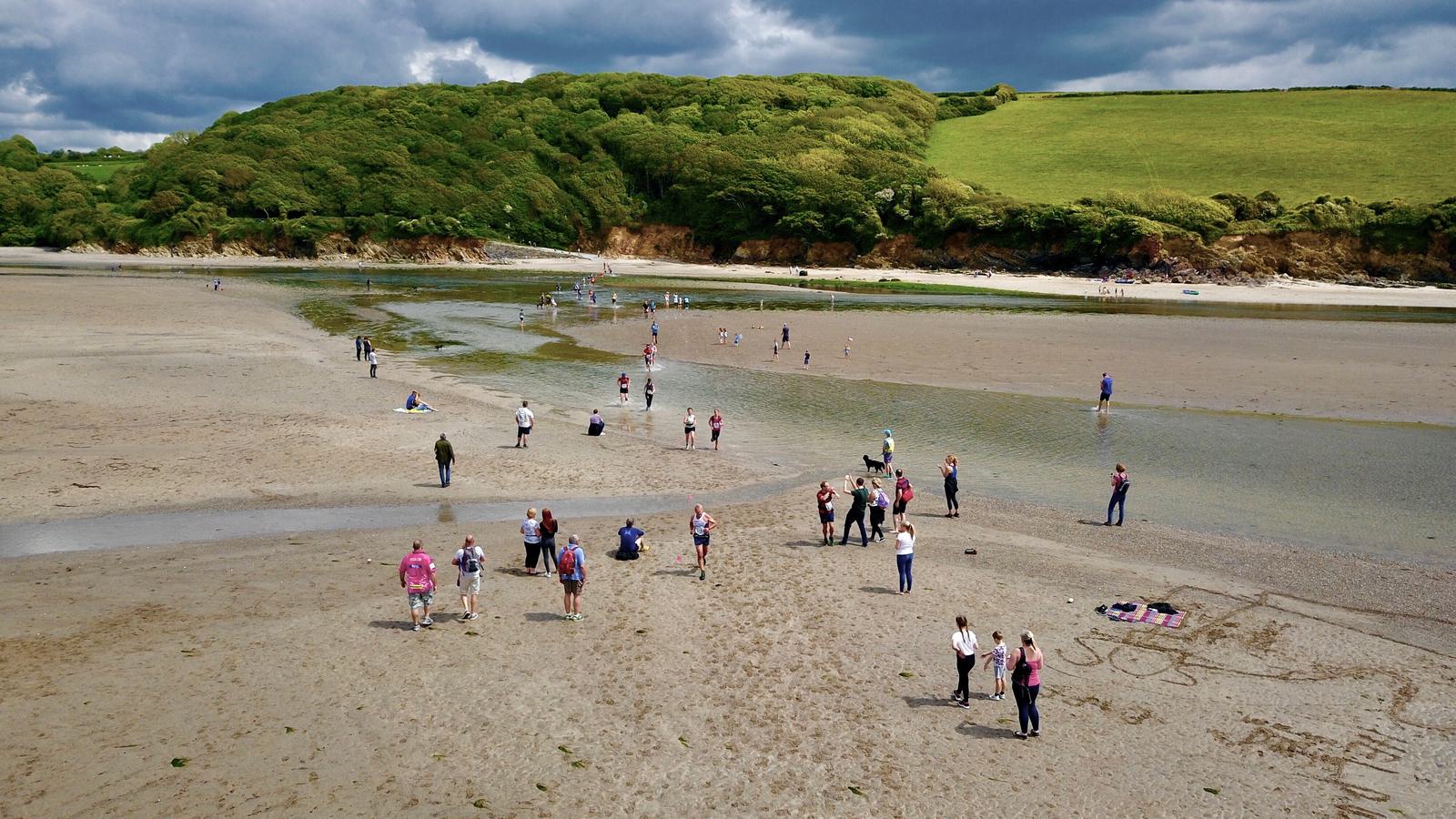 A scenic beach scene with people walking on the sandy shore next to a shallow stream. Green hills and lush trees are visible in the background under a partly cloudy sky.