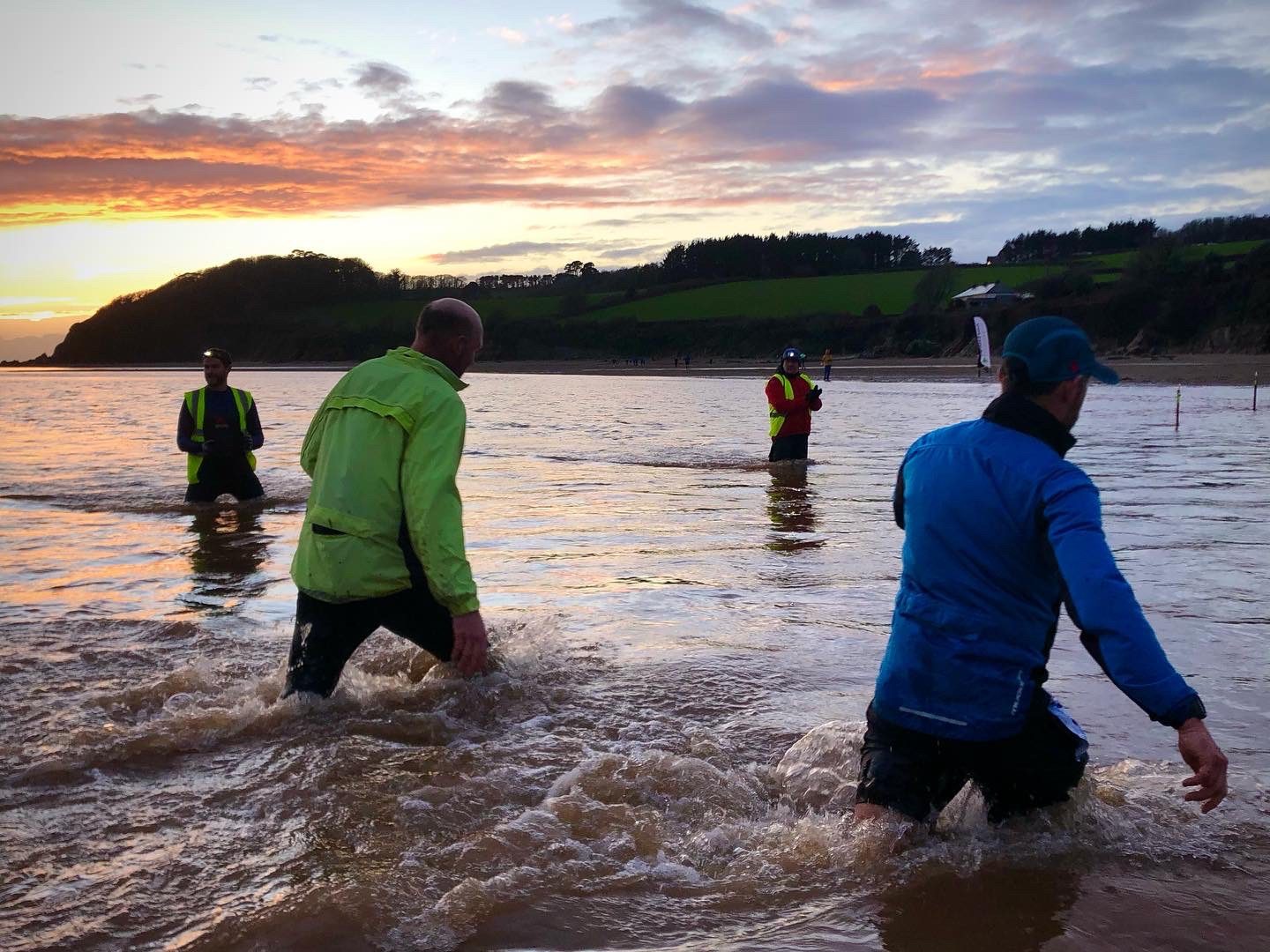 Three people, wearing brightly colored jackets, wade through shallow water at a beach during sunset. A fourth person takes a photo from further back in the water. The background features a hilly landscape with trees, and the sky is painted with orange and blue hues.