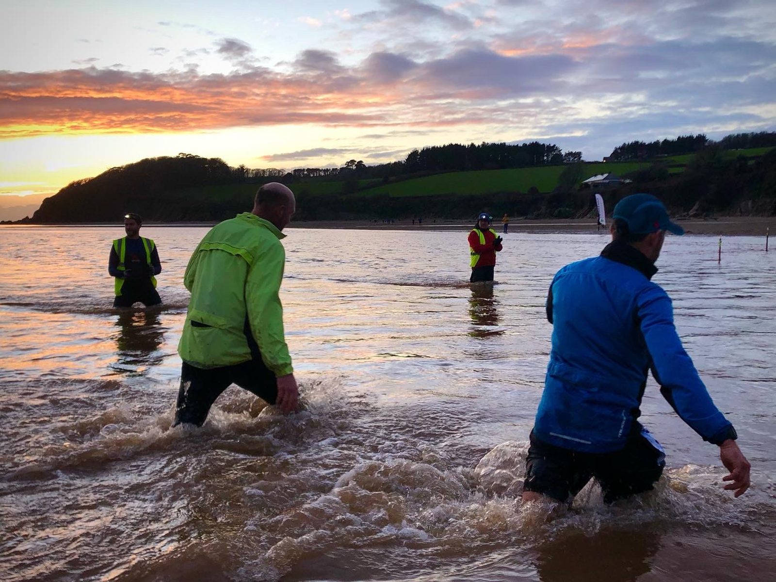Three people, wearing brightly colored jackets, wade through shallow water at a beach during sunset. A fourth person takes a photo from further back in the water. The background features a hilly landscape with trees, and the sky is painted with orange and blue hues.