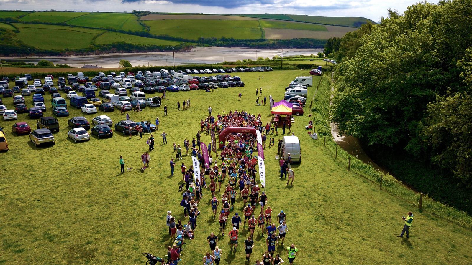 Aerial view of a large group of runners starting a race on a grassy field. A canopy marks the starting line. Cars are parked on one side, and trees border the other. The landscape includes rolling hills and a river in the background.