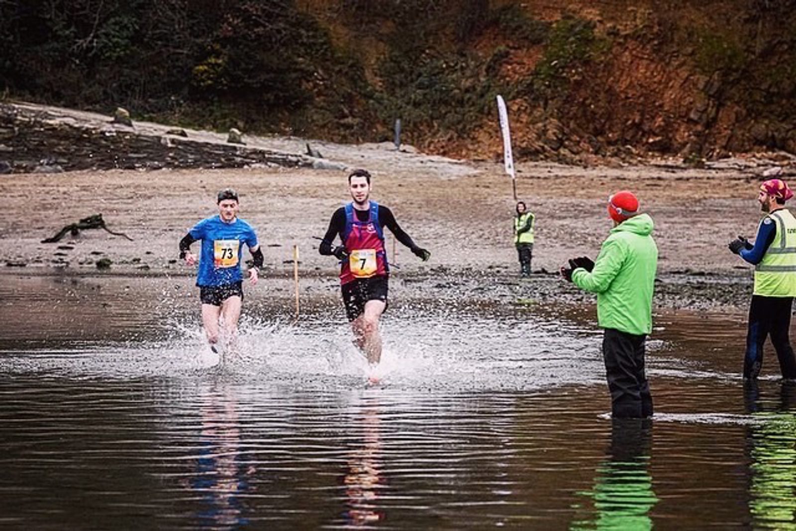 Two runners wearing race bibs numbered 73 and 71 dash through shallow water, kicking up splashes, as part of a race. Officials in bright jackets and one with a camera stand on the shore, with rocky terrain and a wooded backdrop in the background.