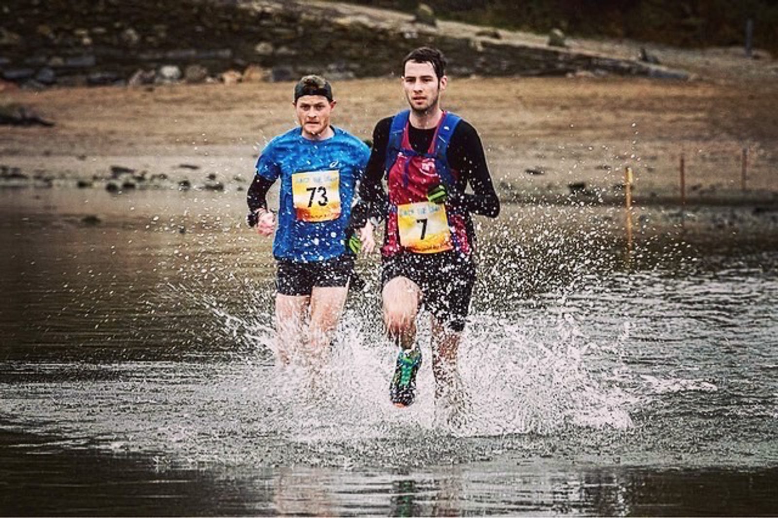 Two runners, each wearing race bibs numbered 73 and 71, are running through a shallow body of water. The runner in front, wearing blue and black, is splashing water as they move forward, with a rocky shoreline visible in the background.