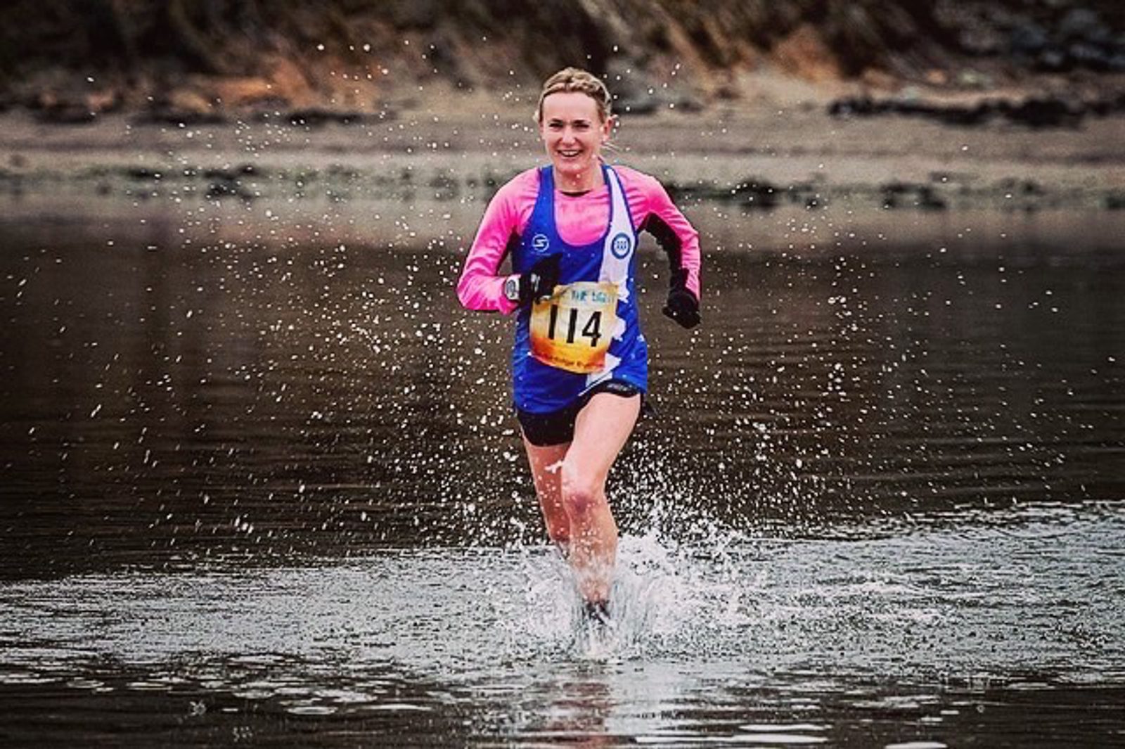 A woman with a race bib numbered 114, wearing a blue vest, pink long-sleeve shirt, and black gloves, runs through shallow water with a smile on her face. The background is blurred, suggesting motion, and consists of a rocky shoreline.
