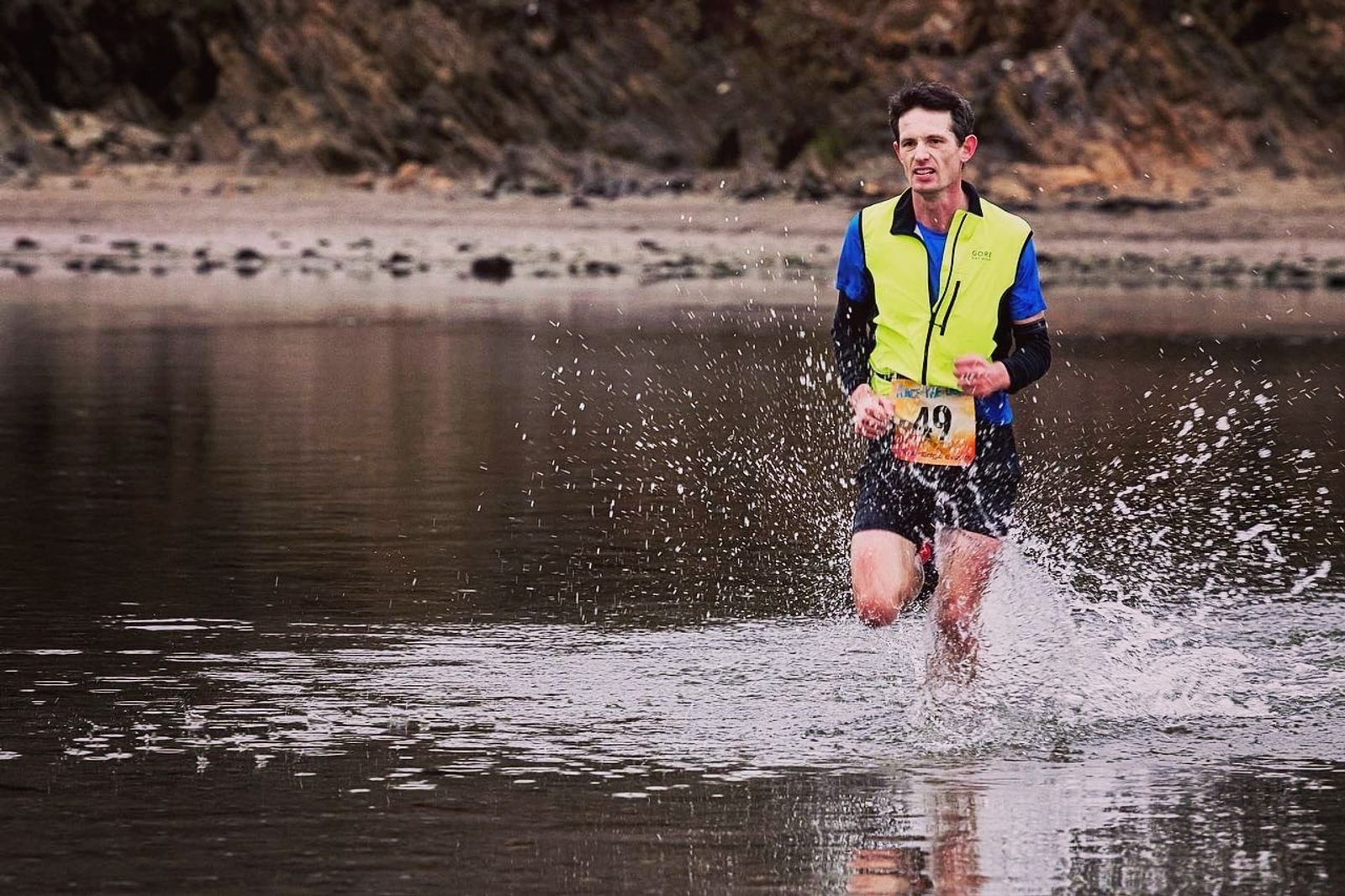 A man in athletic gear runs through a shallow body of water, creating splashes around him. He wears a bright yellow and blue vest with a number tag pinned to it. The background features a rocky, earthy terrain. The expression on his face shows determination.