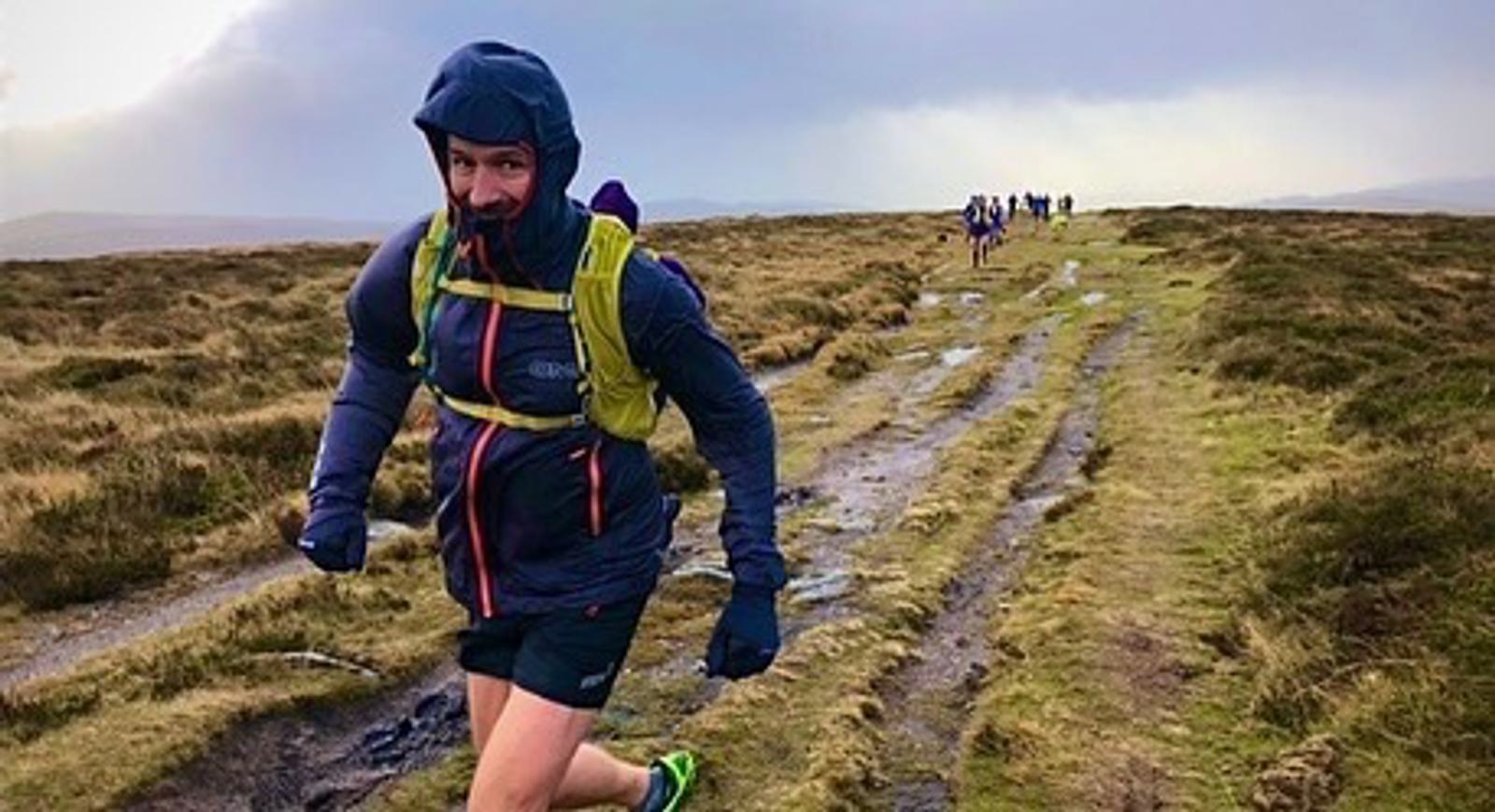 A person wearing outdoor gear, including a hooded jacket and gloves, is running on a muddy trail in a hilly, grassy area. The sky is overcast, and several other runners are visible in the distance on the trail.