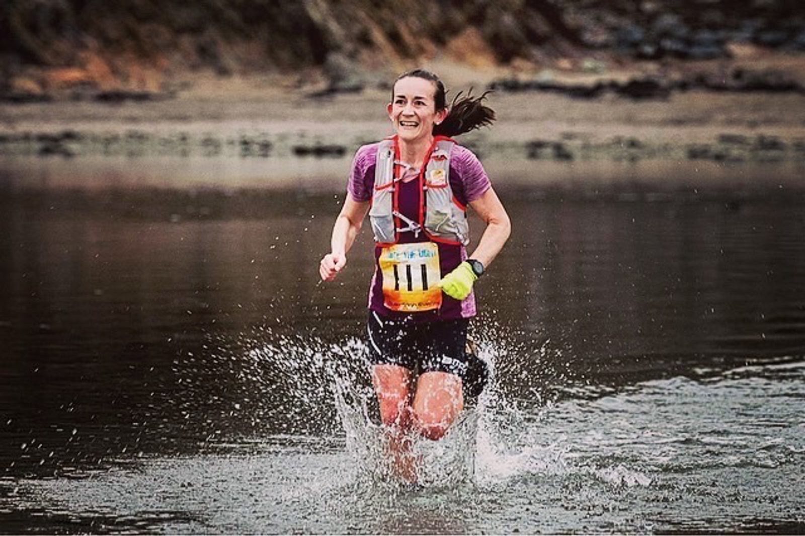 A woman with a race bib numbered 1411 is running through a shallow body of water, splashing around her. She wears a purple shirt, dark shorts, and a hydration vest. She is smiling, with her hair pulled back, and the background shows a rocky, natural landscape.