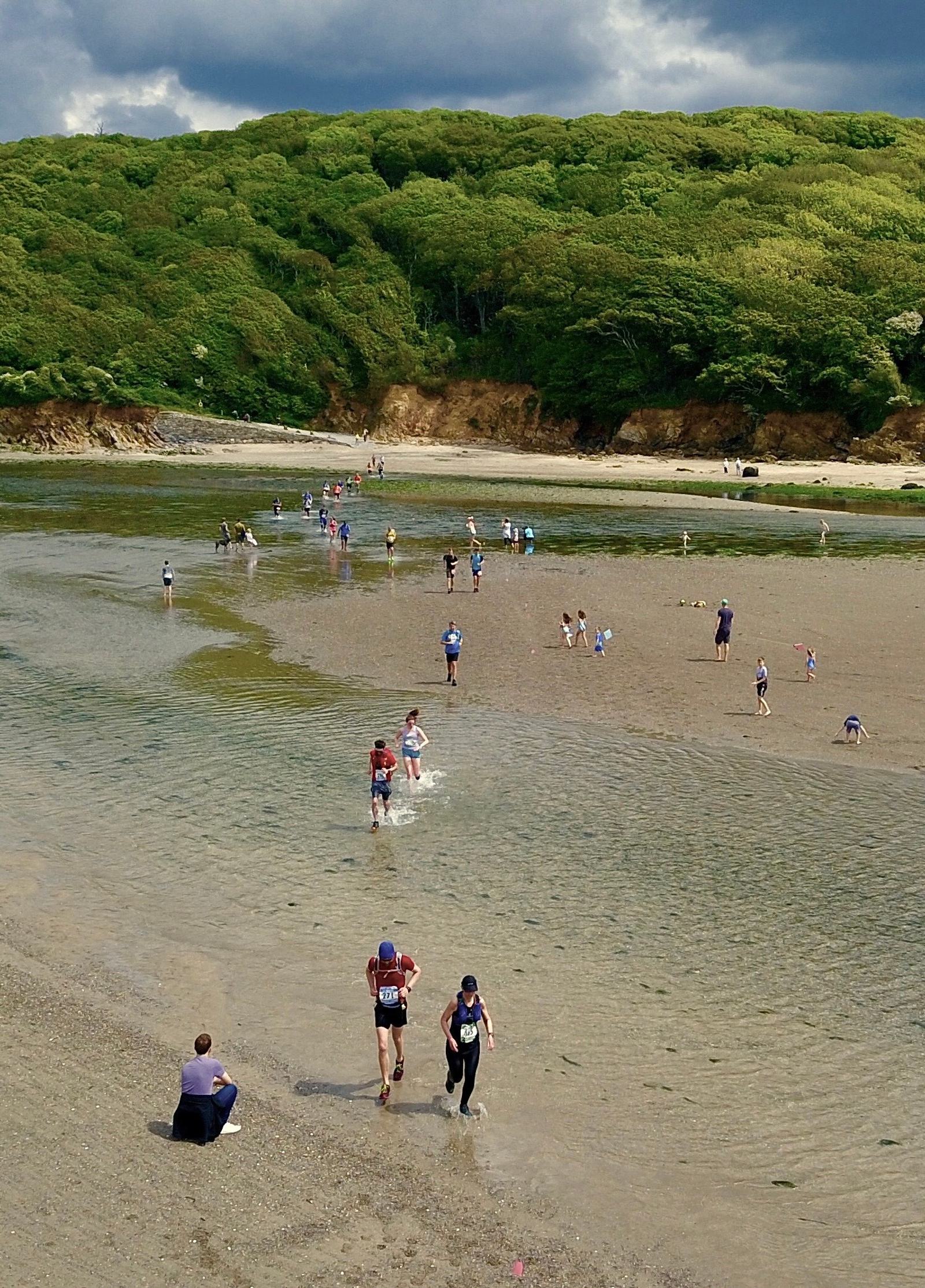 People are running and walking along a beach with a shallow stream, surrounded by lush green trees on a hillside. Some individuals are seated on the shore, while others wade through the water under a cloudy sky.